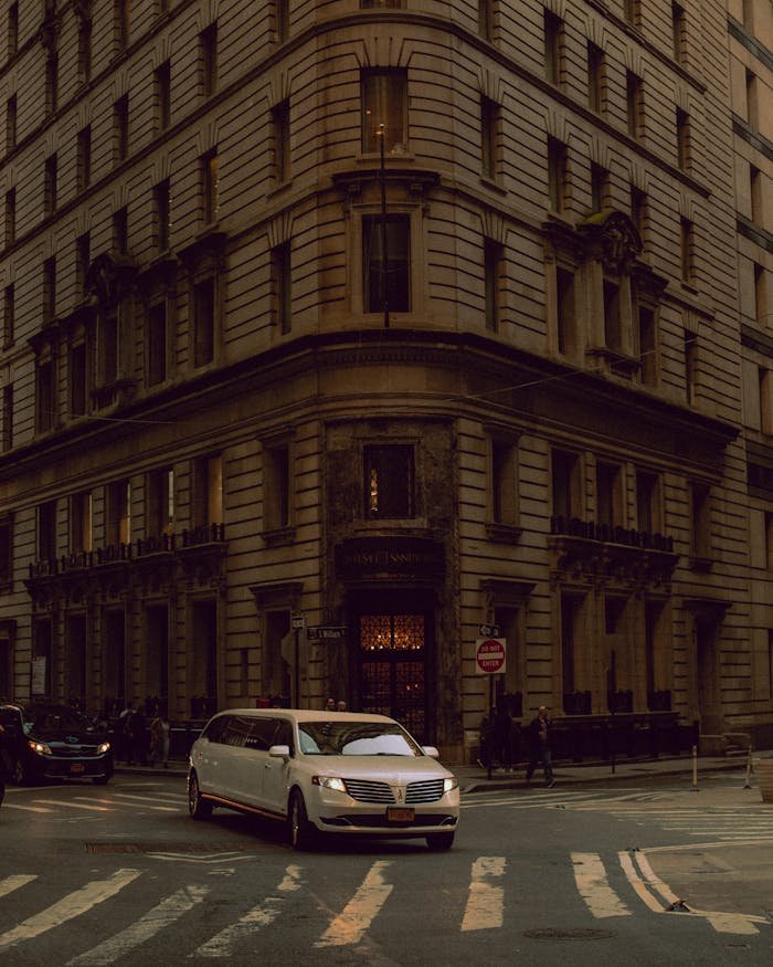 A stylish limousine navigates a bustling city street corner surrounded by towering architecture.