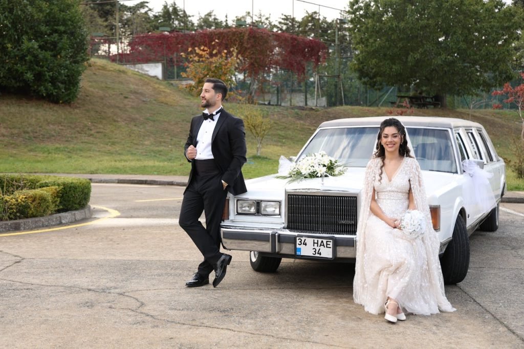 Bride and groom pose with a vintage limousine in a beautiful outdoor setting.